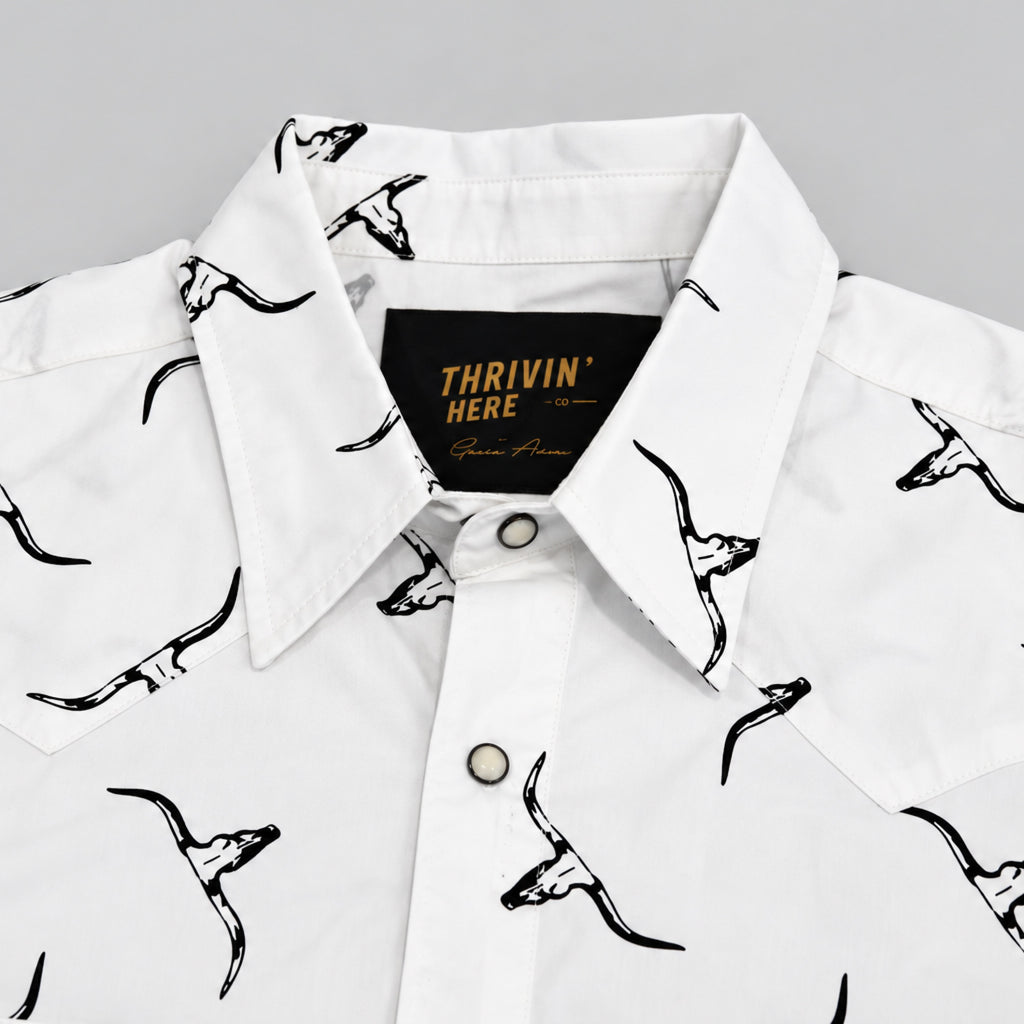 White short-sleeve button-up shirt with a black graphic print of stylized skulls on the front and inside the collar, featuring a black neck label with gold text that reads 'THRIVIN HERE'.