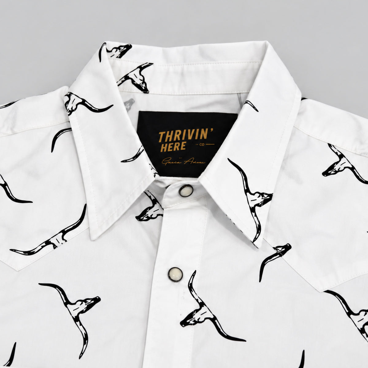 White short-sleeve button-up shirt with a black graphic print of stylized skulls on the front and inside the collar, featuring a black neck label with gold text that reads 'THRIVIN HERE'.