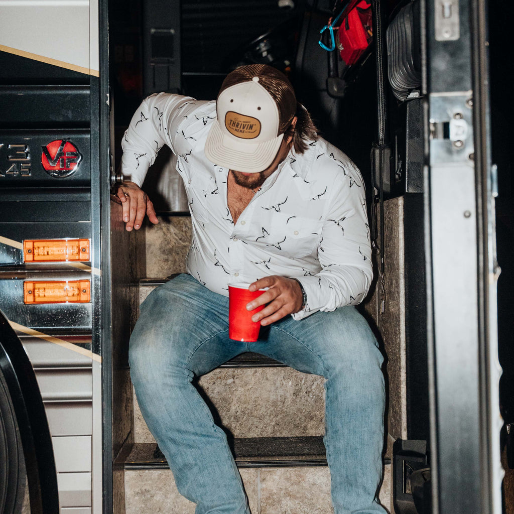Gavinwearing a white patterned button-down shirt, blue jeans, and a beige cap with a brown leather patch sits on the steps of a bus, holding a red cup.
