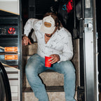 Gavinwearing a white patterned button-down shirt, blue jeans, and a beige cap with a brown leather patch sits on the steps of a bus, holding a red cup.