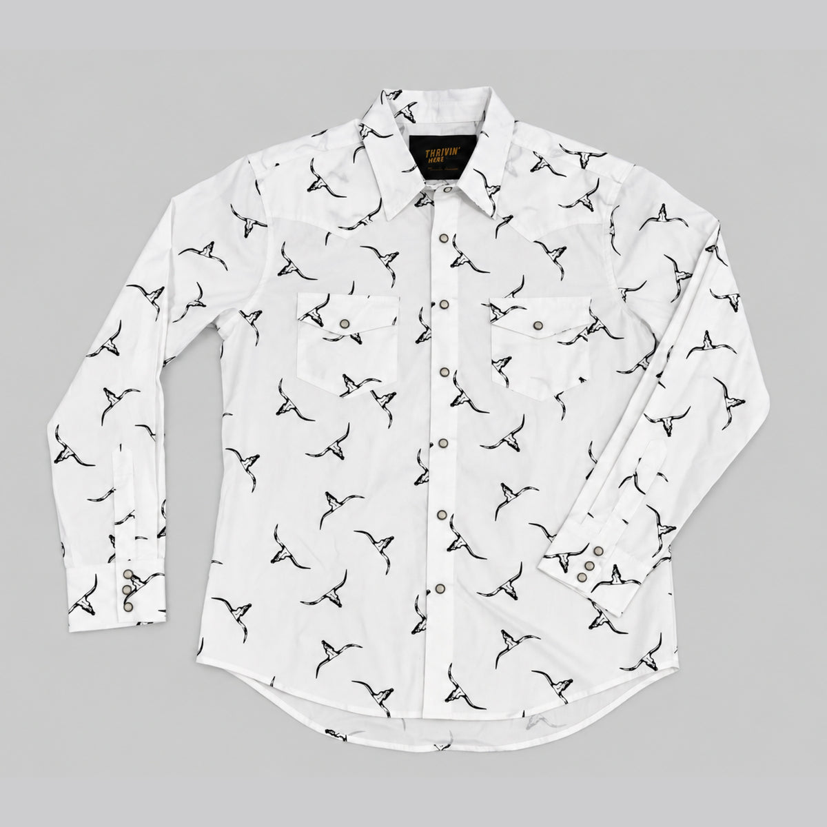 A white long-sleeve button-up shirt with an all-over black bird print, laid flat on a neutral gray background.