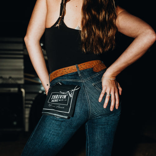 A woman wearing blue jeans with a black patch pocket on the back, styled with a brown belt and a black camisole. The patch pocket features a white logo that reads 'THRIVIN HERE'.